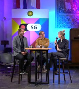 Ewout Meijer, Elwin Hofman and Els Aarts sit around a table. Els Aarts is asking a question, the two others are listening. Behind them is a colourful banner with the logo of Utrecht University and Studium Generale.